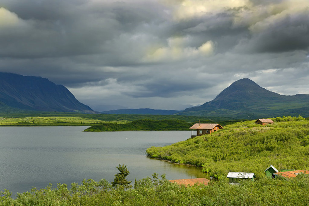 Groene heuvels met hutten aan een meer, met bergen op de achtergrond in Alaska.