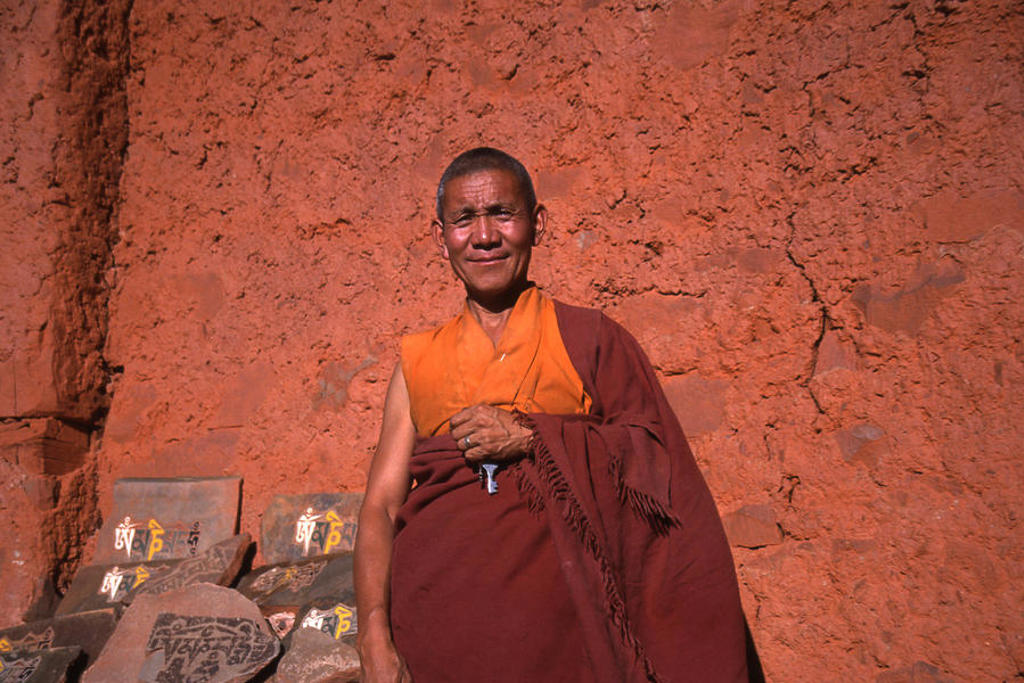 Boeddhistische monnik in Ladakh, India, met gebedsstenen tegen een rood lemen muur.