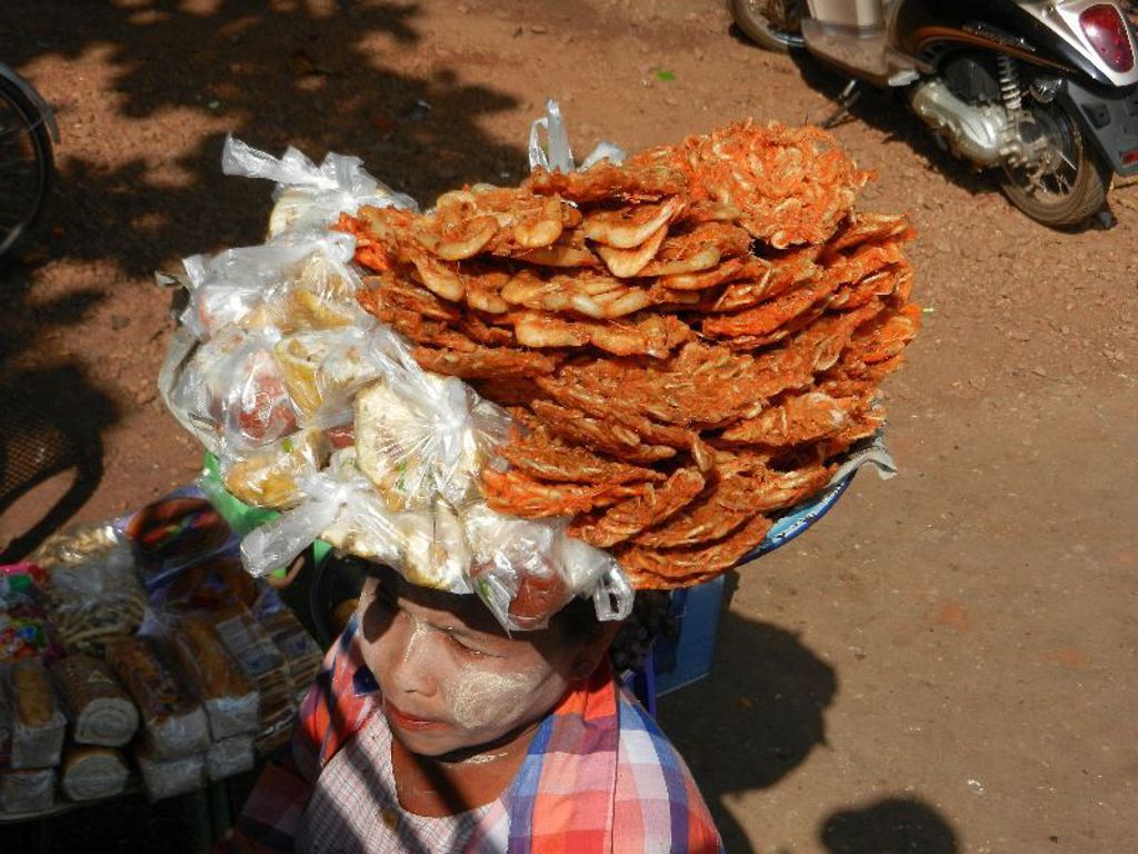 Marktvrouw in Myanmar met thanaka op haar gezicht en een stapel snacks op haar hoofd.