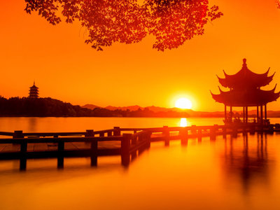Zonsondergang bij een Chinese pagode aan een meer, met houten pier en silhouetten in warm oranje licht.
