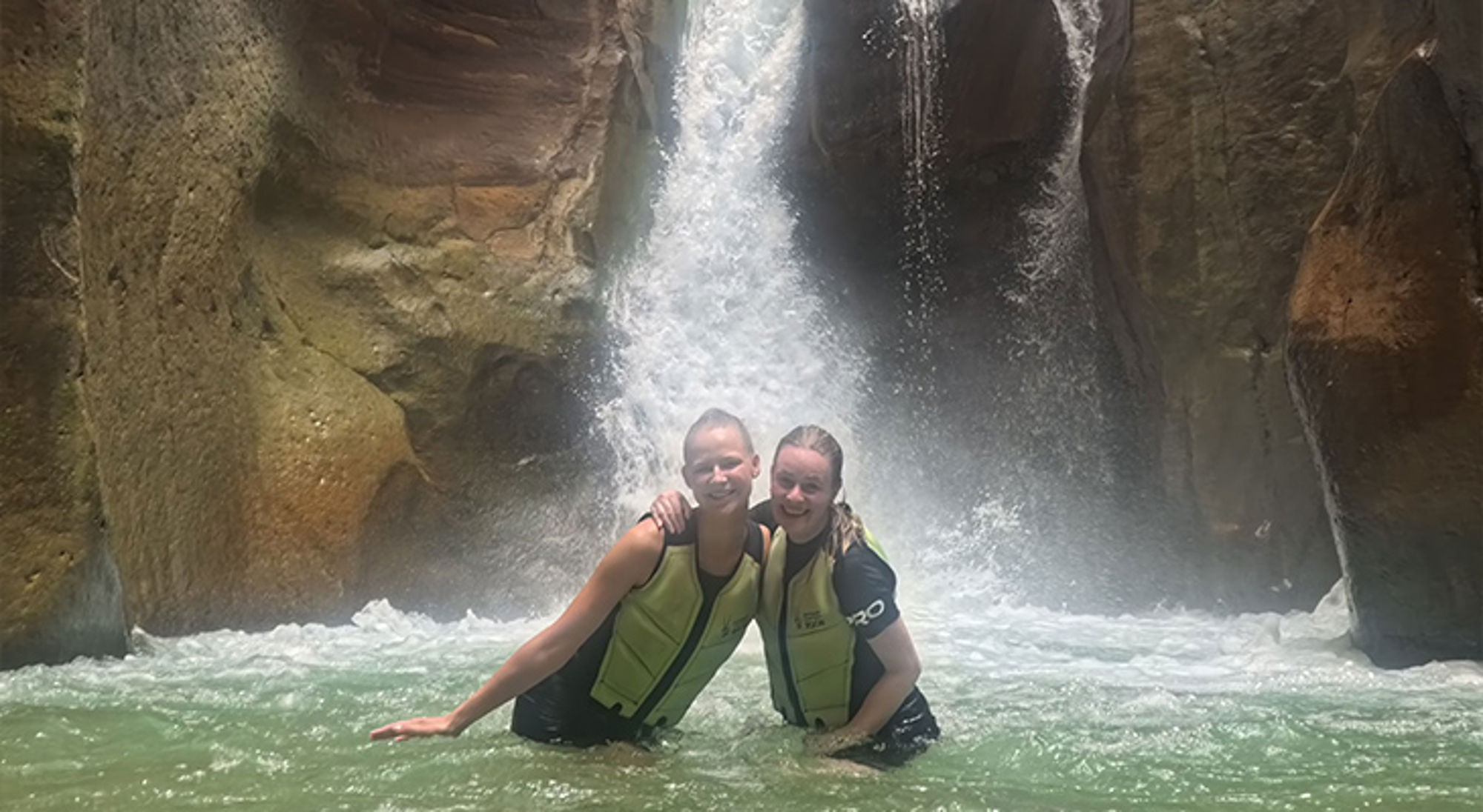 Twee reizigers in zwemvesten poseren in een rivier bij een waterval in een smalle rotskloof.
