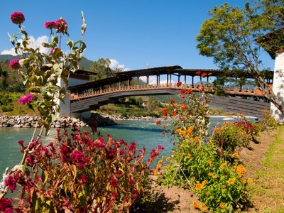 Overdekte houten brug over een rivier in Bhutan, omringd door bloemen en bergen.
