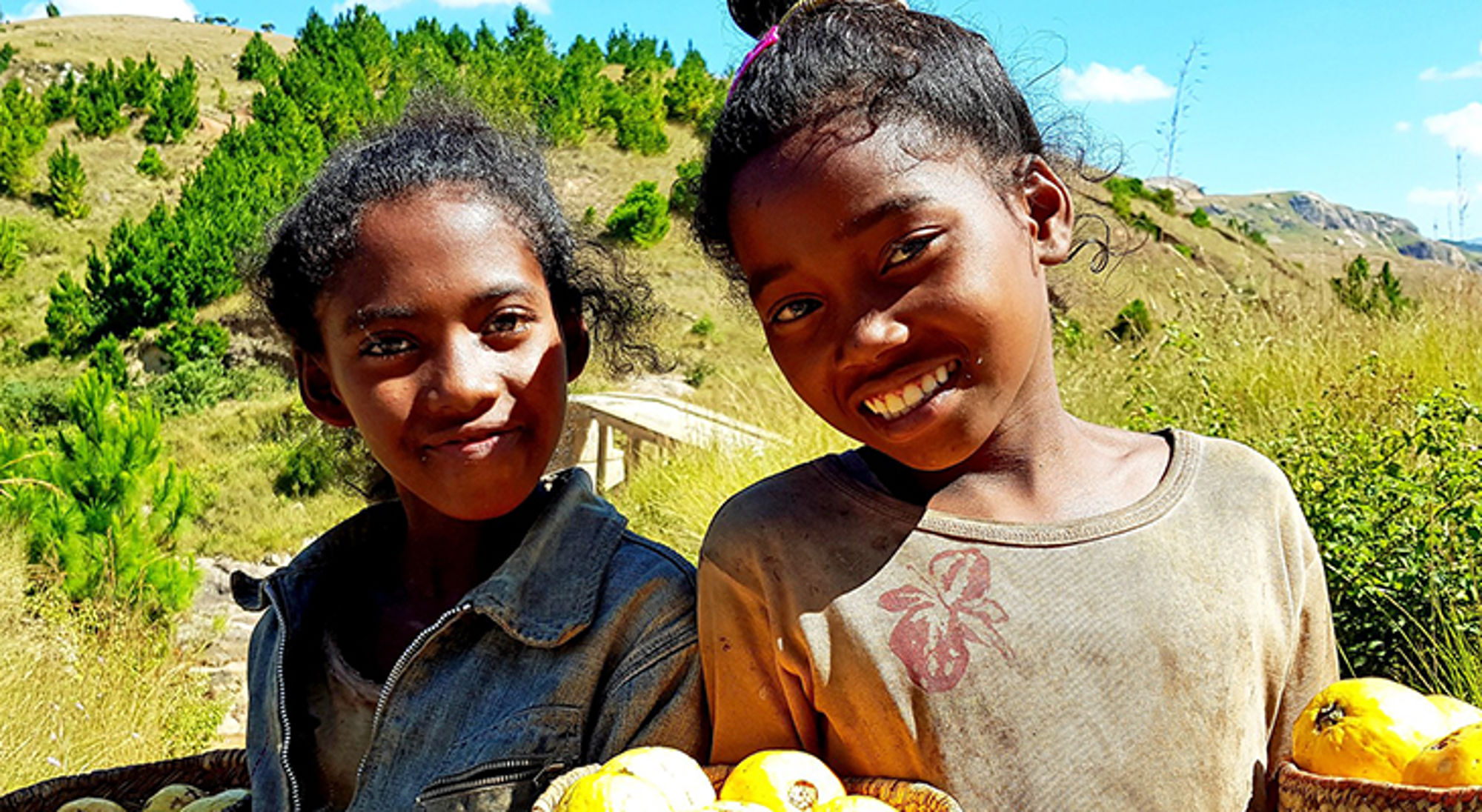 Twee lachende meisjes met manden vol fruit langs een bergpad in een groen heuvellandschap.