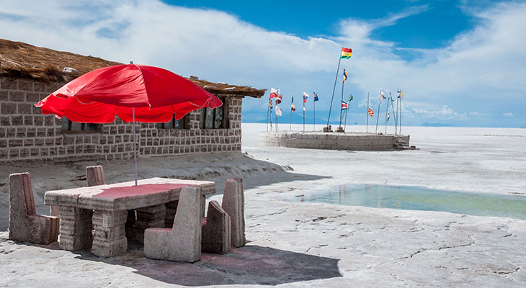 Rode parasol boven stenen tafel en banken aan de zoutvlakte, met vlaggen op de achtergrond.