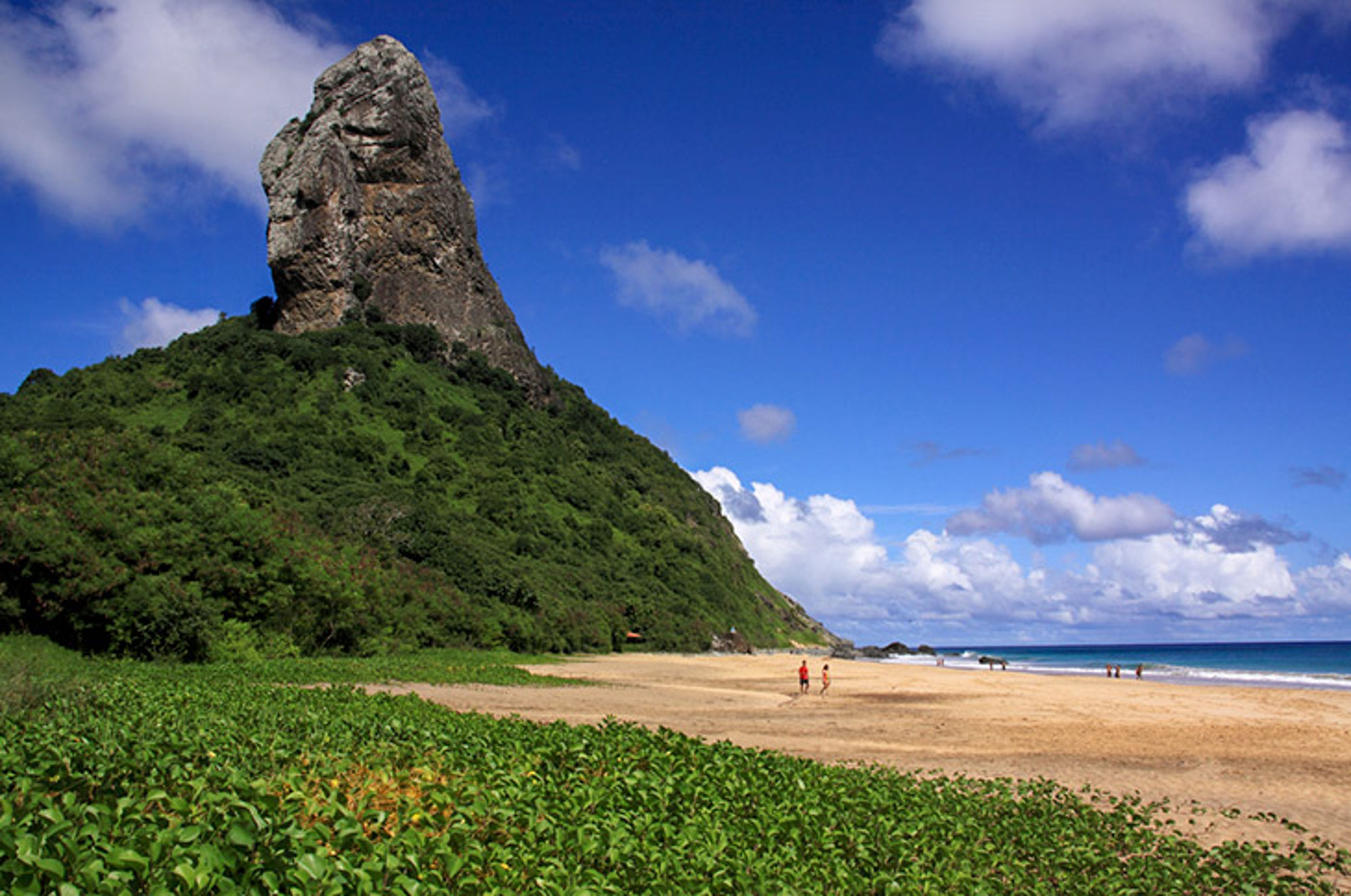 Morro do Pico rotsformatie bij Praia do Sancho op Fernando de Noronha, met strand, zee en blauwe lucht.