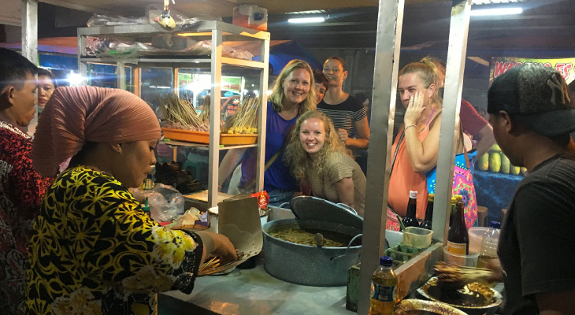 bij eetkraampje in azie op singlereis Straatkraam op een avondmarkt waar saté wordt bereid, met reizigers die toekijken en lachen.