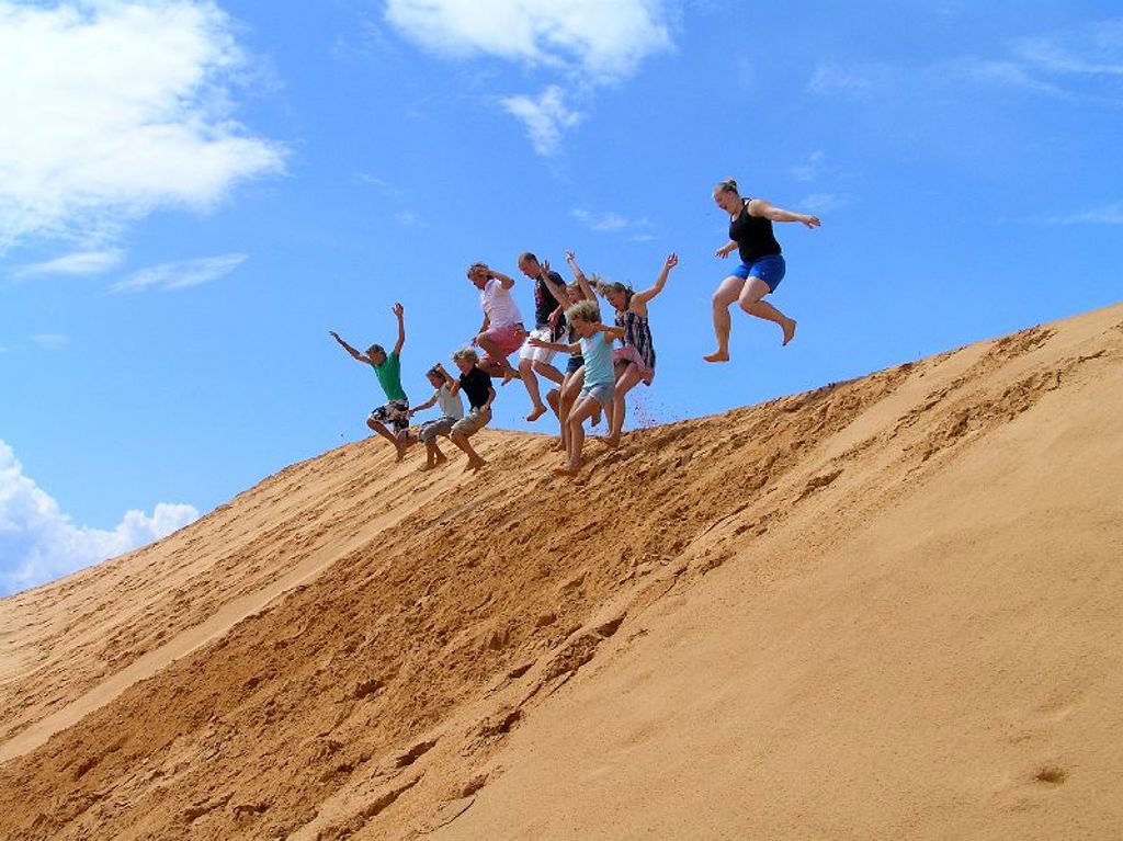 Gezin springt van een zandduin in Vietnam tijdens een familiereis, met blauwe lucht op de achtergrond.