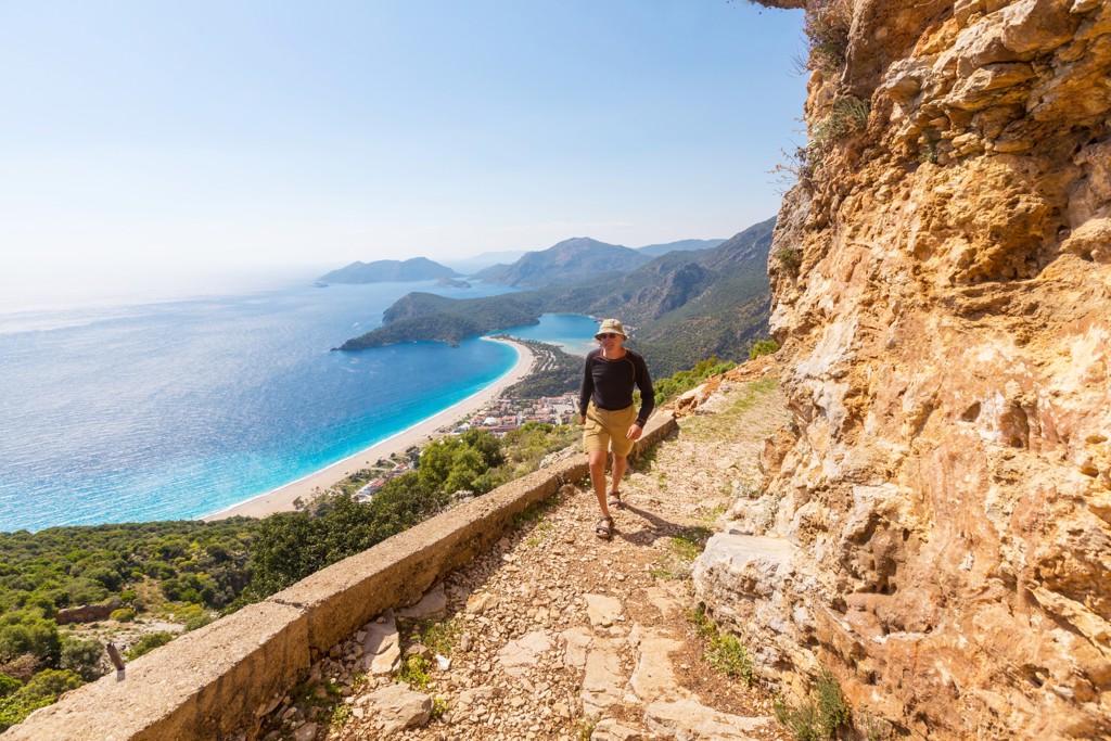 Wandelaar op bergpad langs de Turquoise Kust in Turkije met uitzicht op azuurblauwe baai en strand.