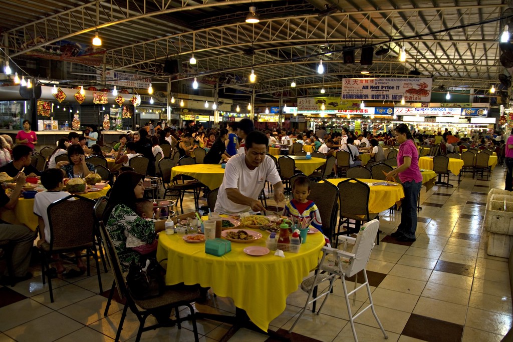 Drukke foodcourt in Maleisisch Borneo met reizigers en locals aan tafels onder felle marktverlichting.