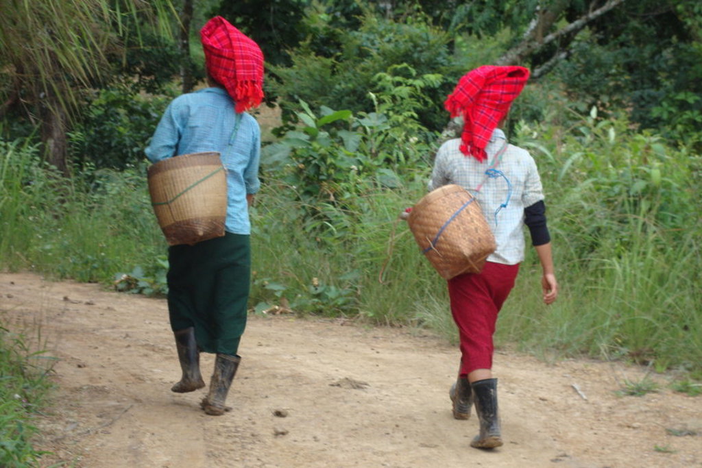 Twee vrouwen met rode hoofddoek en mand op de rug lopen over een zandpad in Myanmar.