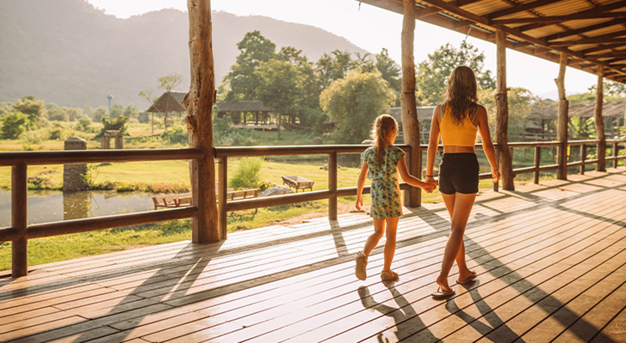 Moeder en dochter wandelen hand in hand over een houten veranda met uitzicht op bergen en natuur.