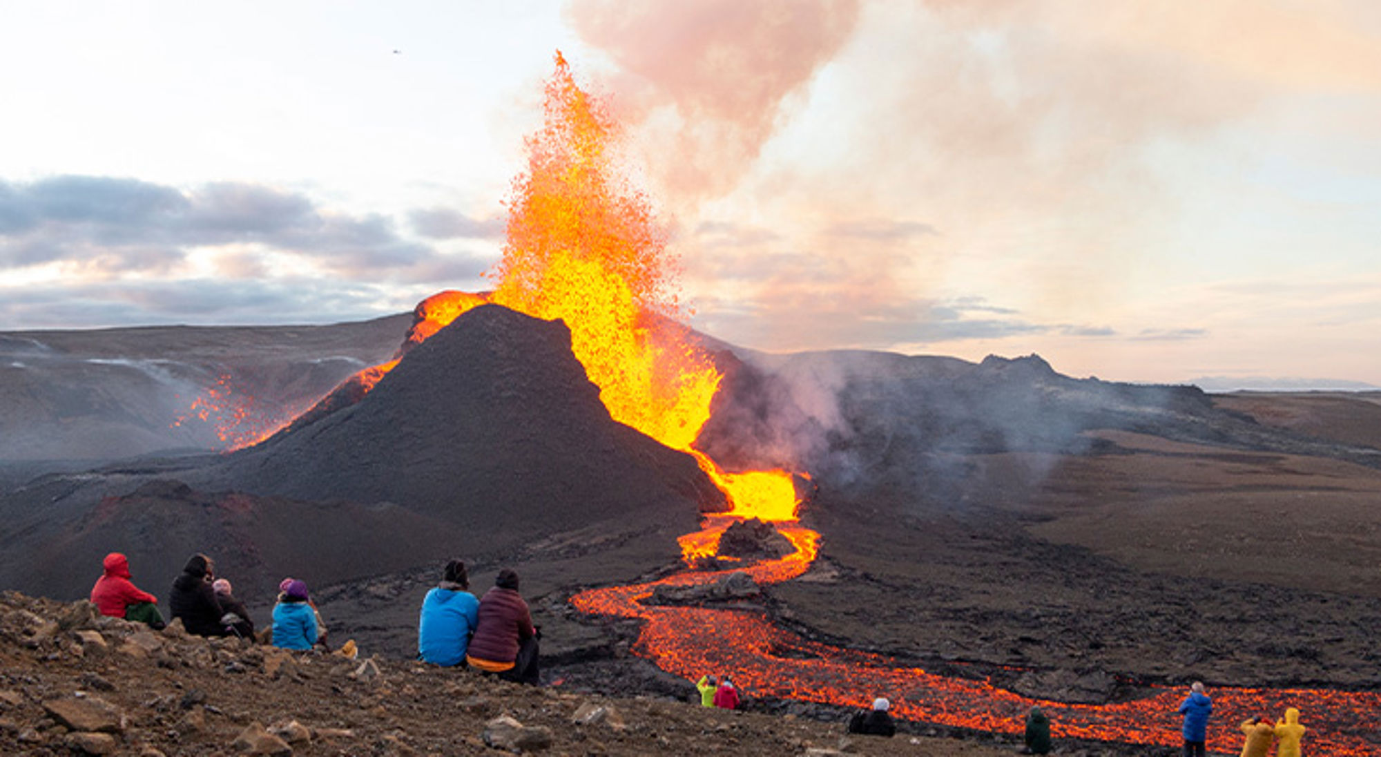 Fagradalsfjall uitbarsting Toeristen kijken naar een vulkaanuitbarsting met lavafontein en lavastroom in een ruig landschap.