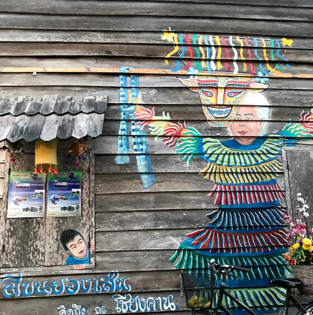 Muurschildering op houten gevel in Thailand met kleurrijk figuur en kind dat uit een raam kijkt.