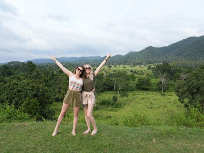 Twee reizigers poseren met opgeheven armen bij een groen berglandschap in Thailand.