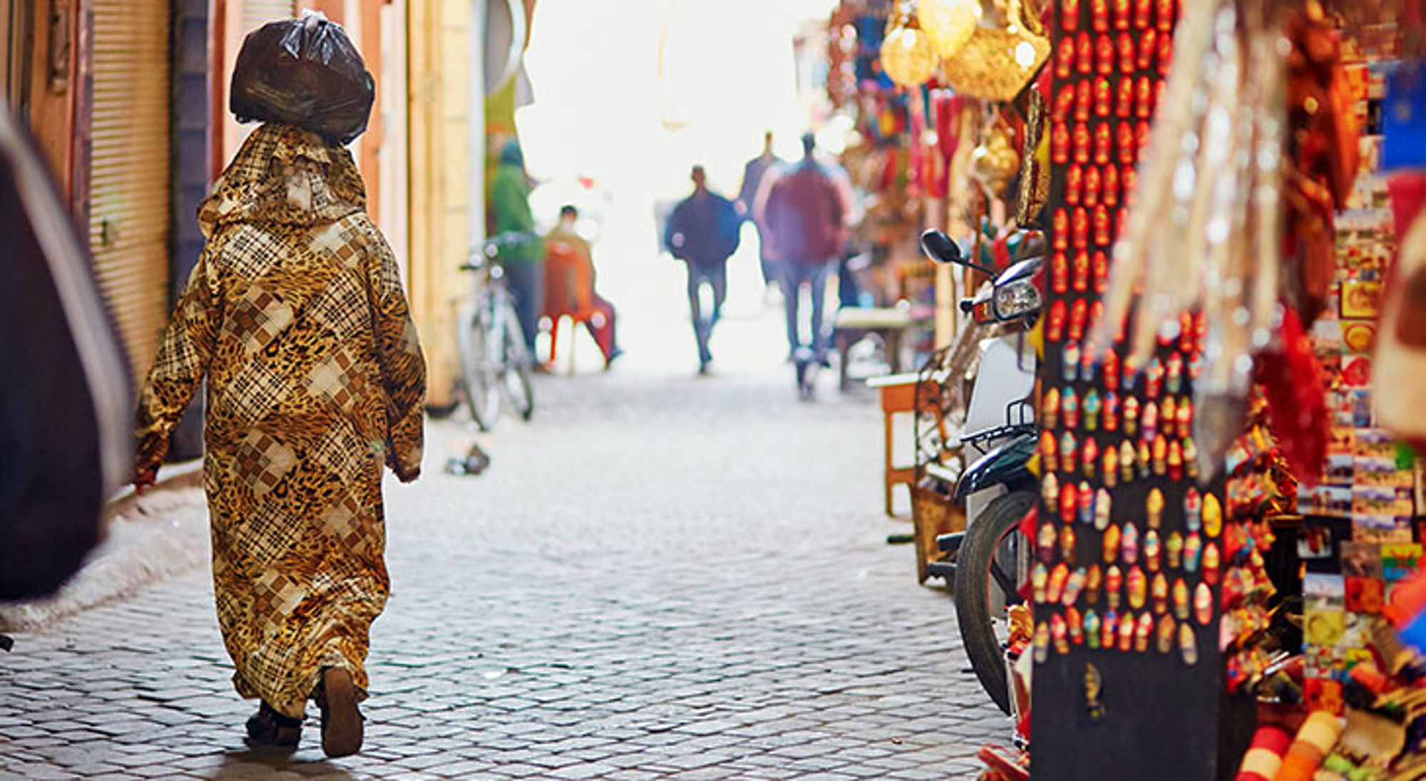 De mooiste stad ter wereld Marrakech Vrouw loopt door smalle straat met marktkraampjes en souvenirs, met een tas op het hoofd.