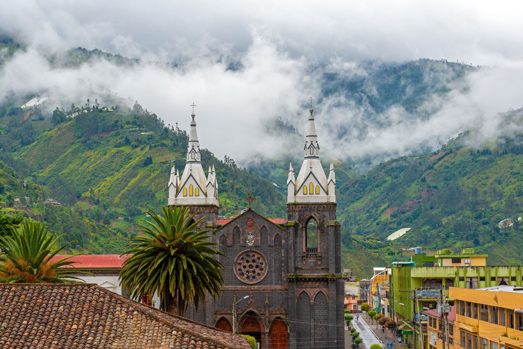 Kerk met twee torens in Baños, Ecuador, met groene bergen en mist op de achtergrond.
