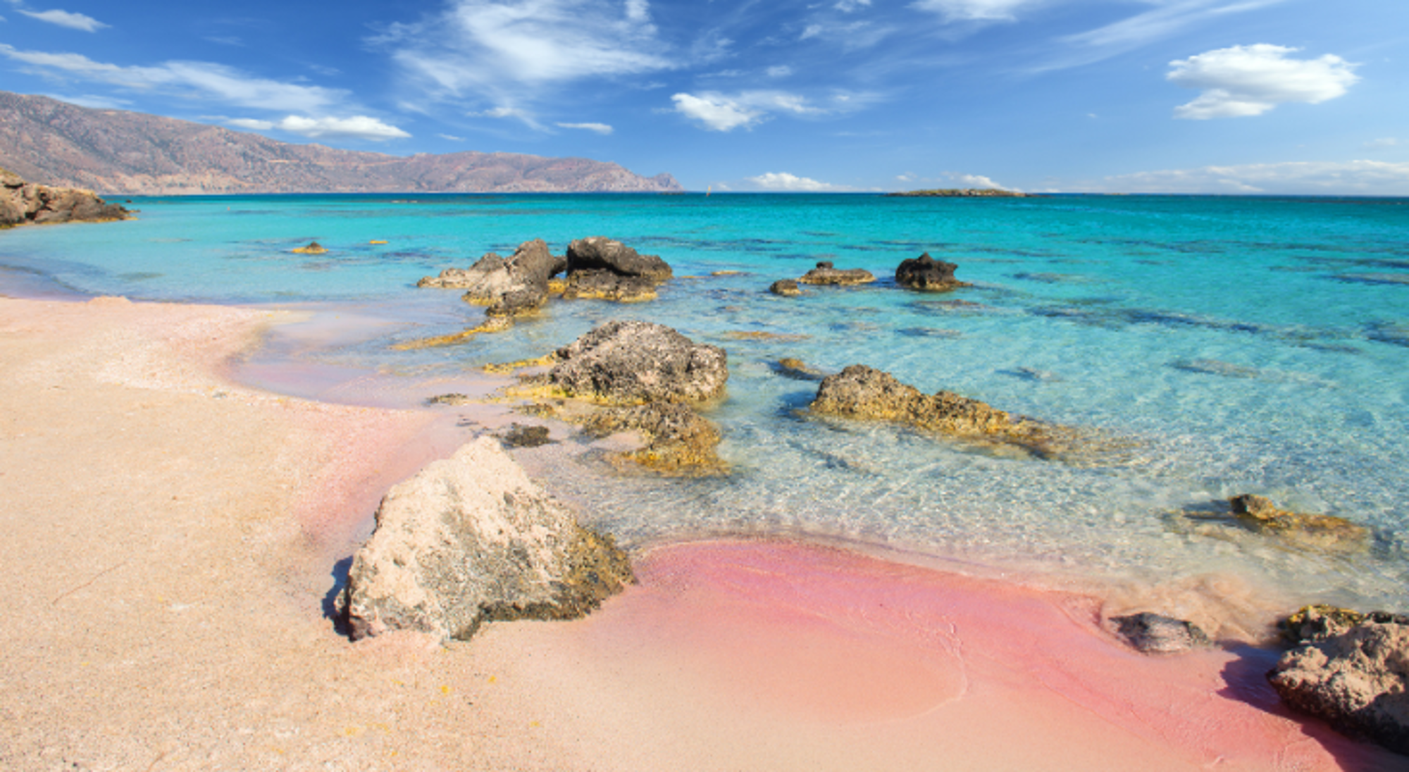 Roze zandstrand met rotsen en helder turquoise zeewater onder een blauwe lucht.