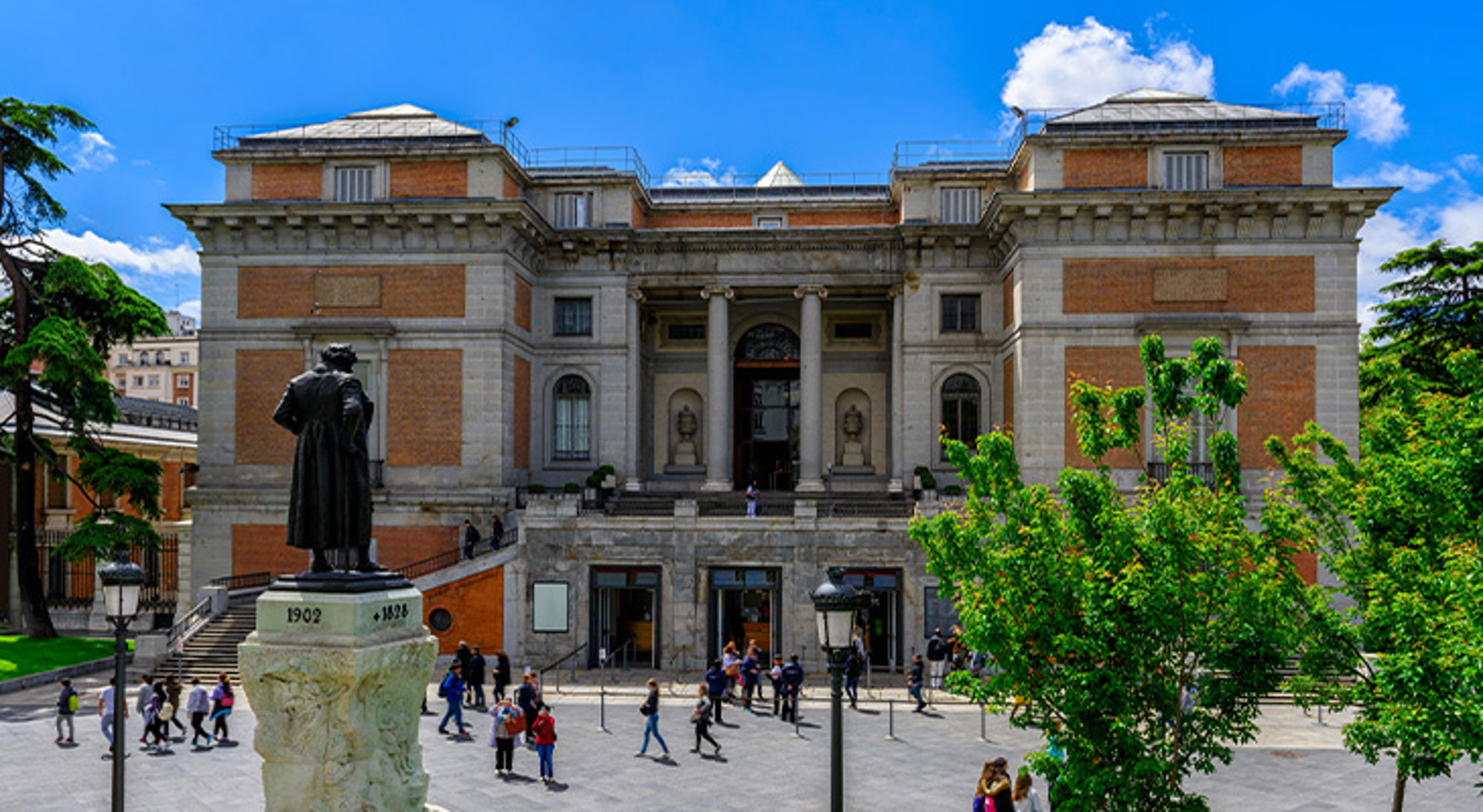 Museo del Prado, Madrid Voorgevel van het Prado Museum in Madrid met bezoekers op het plein en een standbeeld op de voorgrond.
