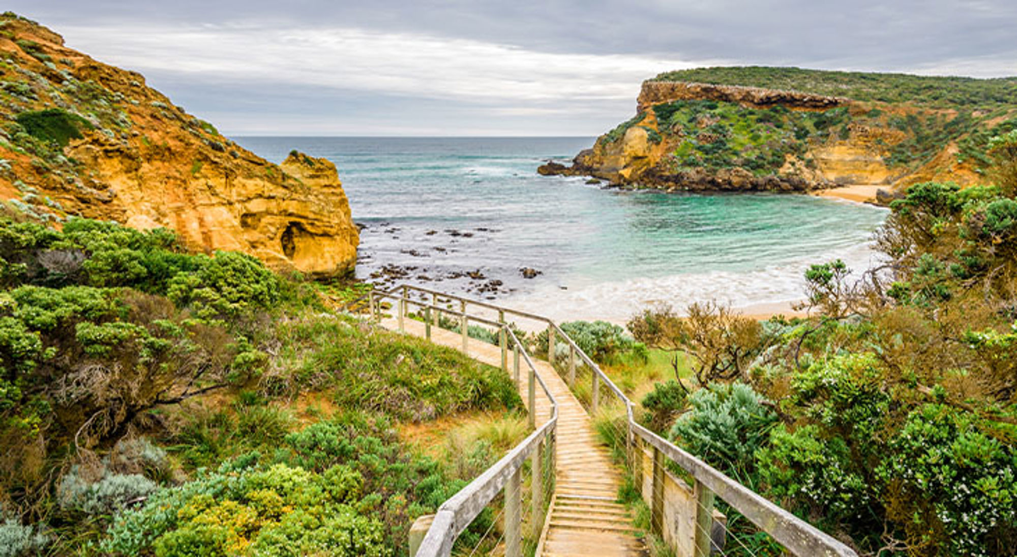 Great Ocean Road Houten wandelpad naar een beschutte baai met zandstrand, rotskliffen en zee onder een bewolkte lucht.