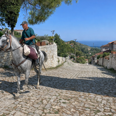 Man te paard in traditioneel dorp in Albanië, met uitzicht op zee en heuvels.