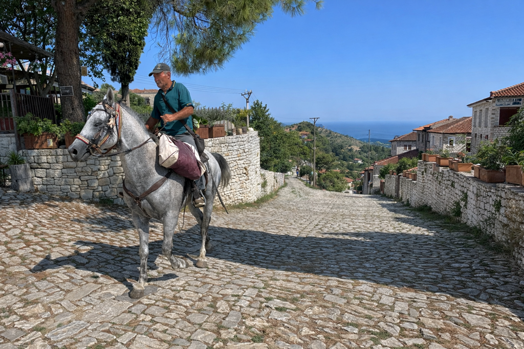 Man te paard in traditioneel dorp in Albanië, met uitzicht op zee en heuvels.