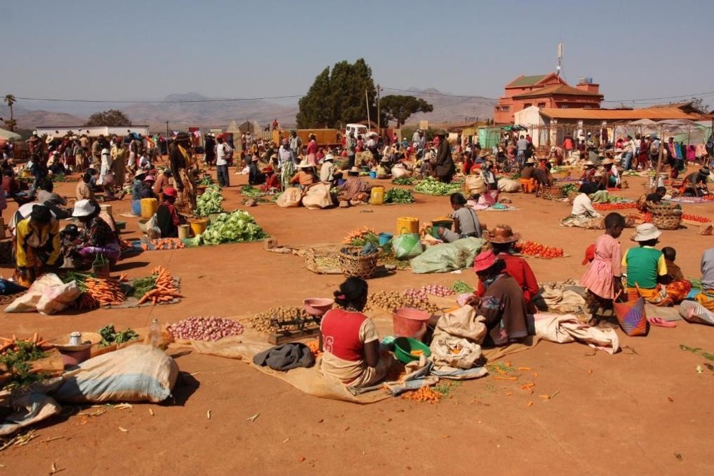 Drukke openluchtmarkt in Madagascar met verkopers en stapels groente op het zand.