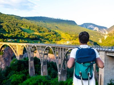 Rugzakreiziger kijkt uit over een hoge boogbrug in een groene bergvallei langs de Route Balkan.