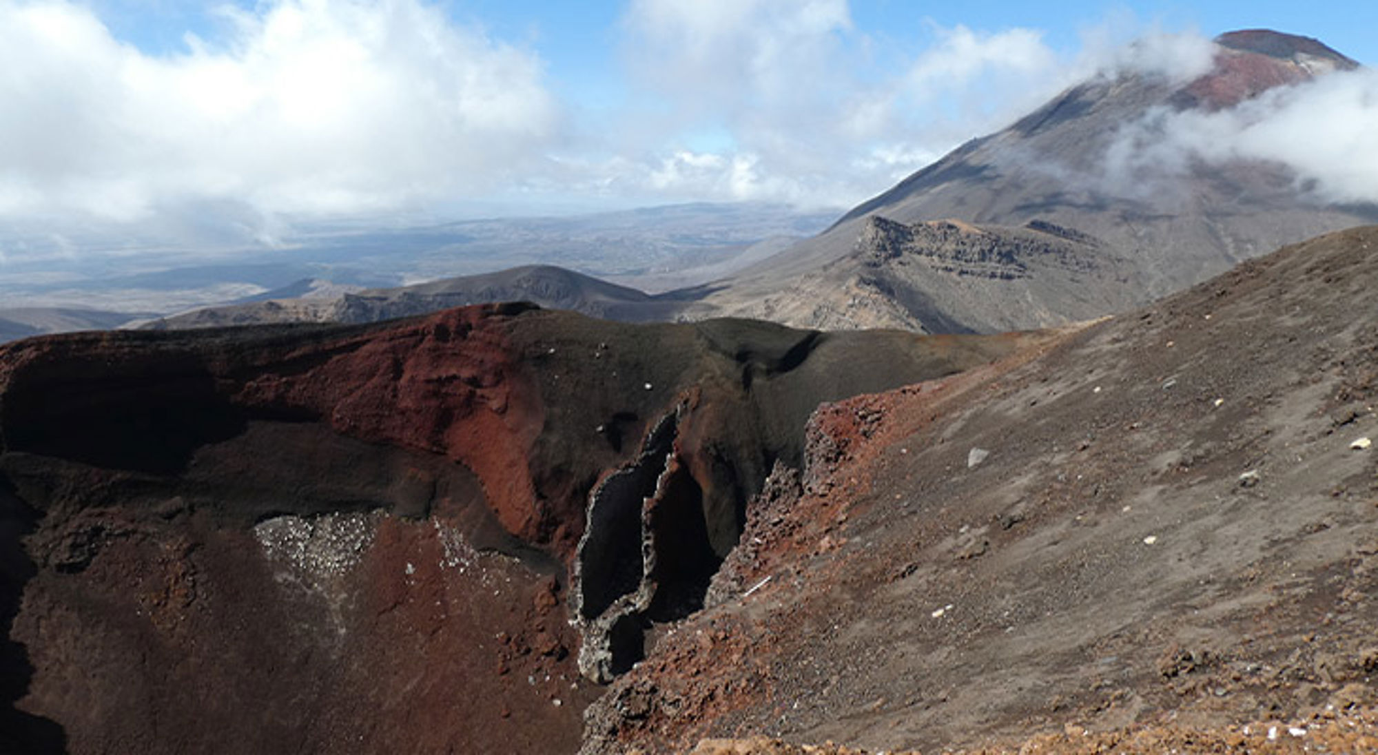 Red Crater Uitzicht op een vulkaankrater met rood en zwart lavagesteente en wolken rond de bergtop.