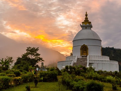 Witte boeddhistische stoepa in Nepal bij zonsondergang, met groene tuin en heuvels op de achtergrond.