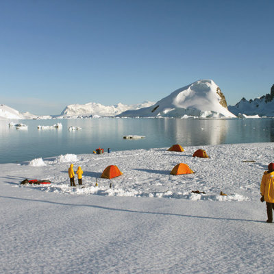 Kampeerplek op het Antarctische ijs met tenten en reizigers, met ijsbergen en bergen op de achtergrond.