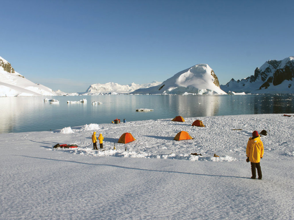 Kampeerplek op het Antarctische ijs met tenten en reizigers, met ijsbergen en bergen op de achtergrond.