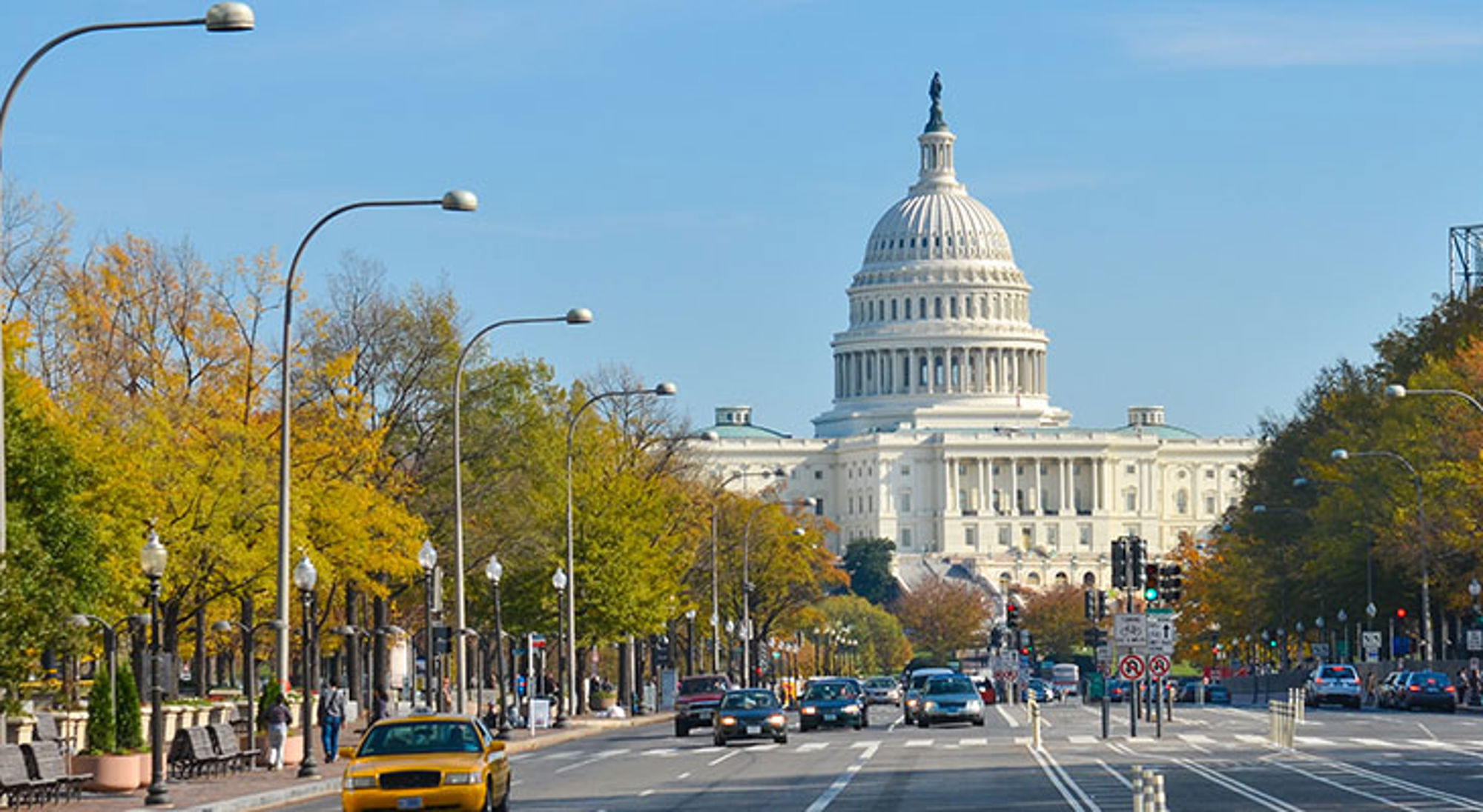Uitzicht op het Capitool in Washington D.C. vanaf een brede straat met verkeer en bomen in herfstkleuren.
