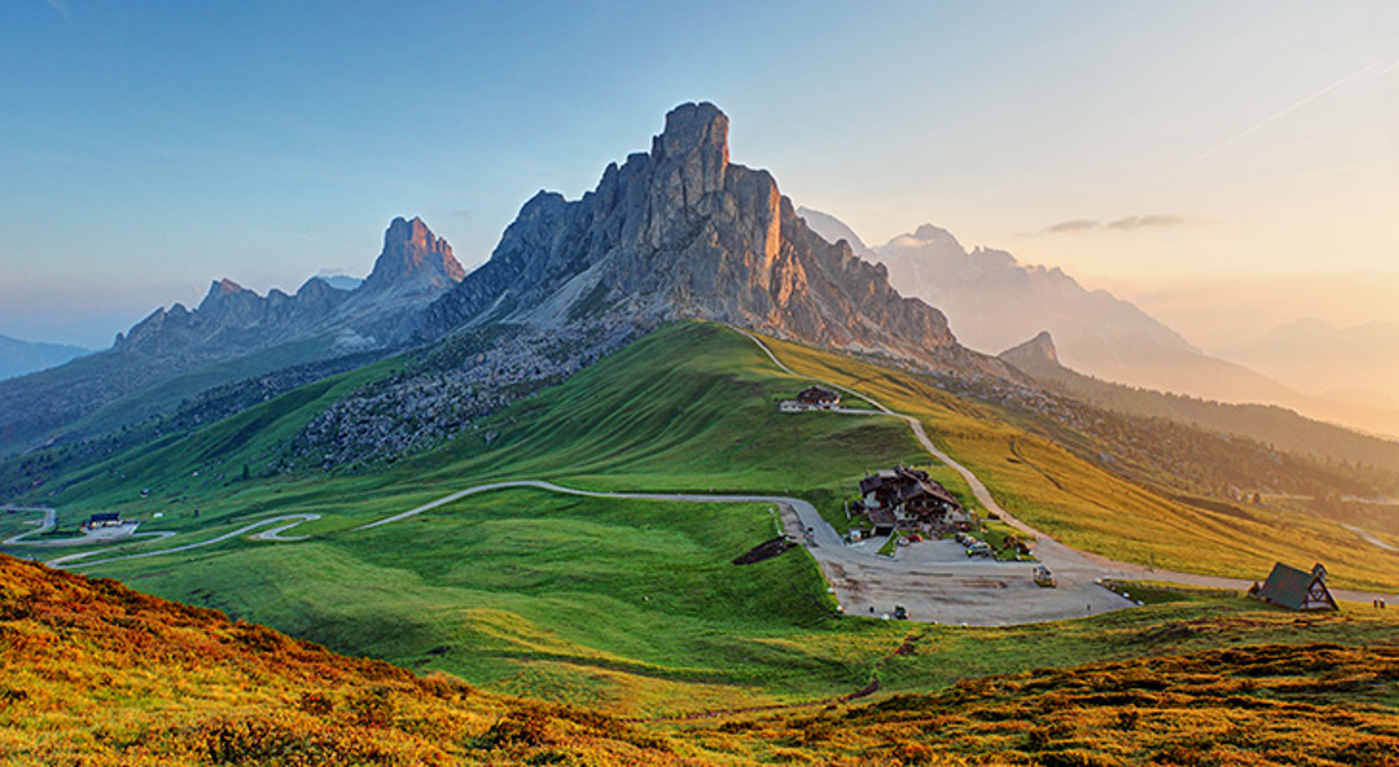 italie-dolomieten Berglandschap in de Dolomieten met groene weiden, berghut en kronkelende weg bij zonsopkomst.