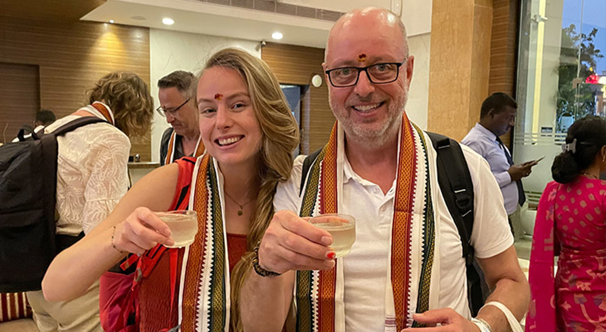 Twee reizigers proosten met een welkomstdrankje in een hotelreceptie.