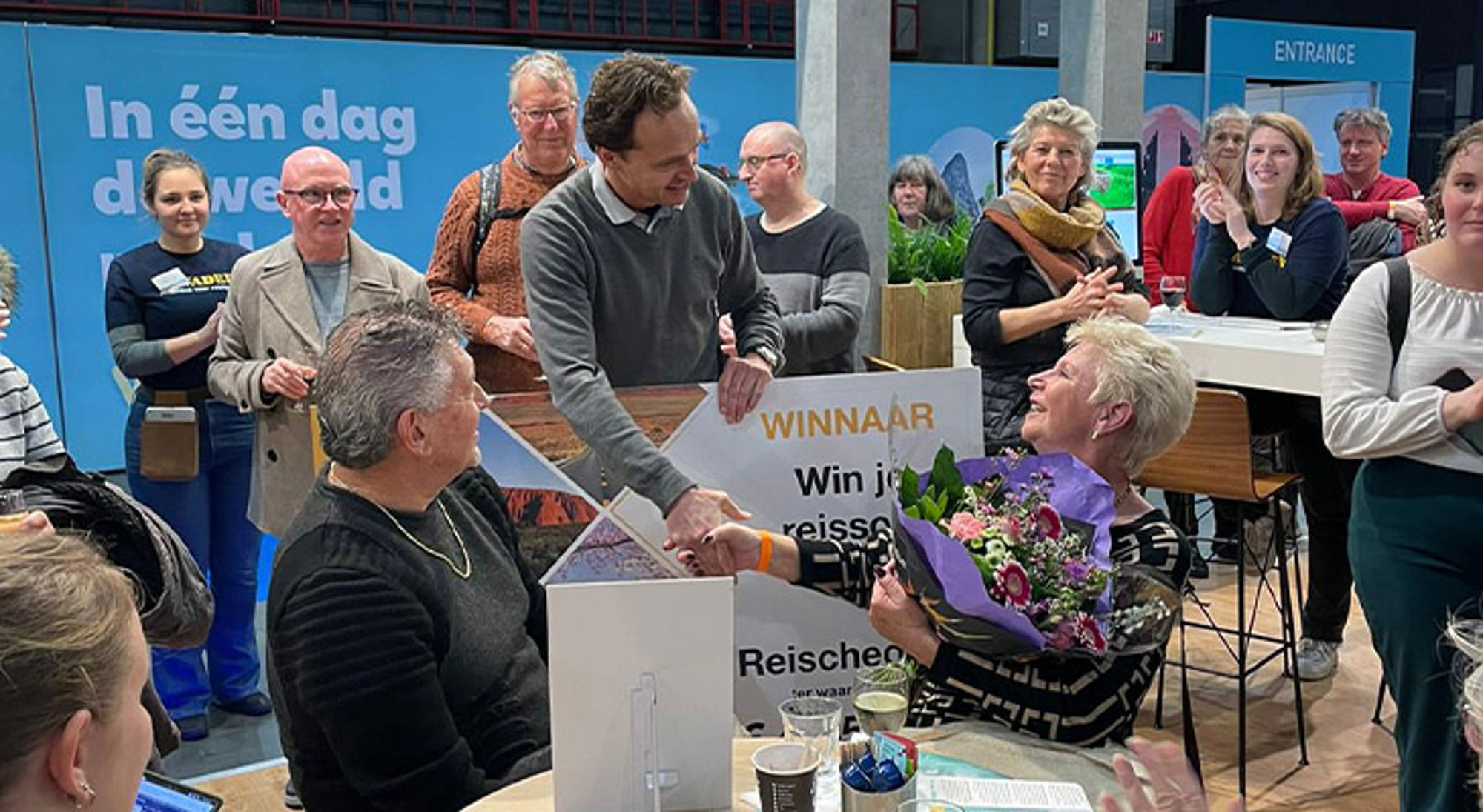 Prijsuitreiking met winnaar die een boeket bloemen ontvangt tijdens een reisbeursactie.
