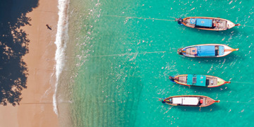strand in Thailand met turquoise zee en vier longtailboten langs de kust.