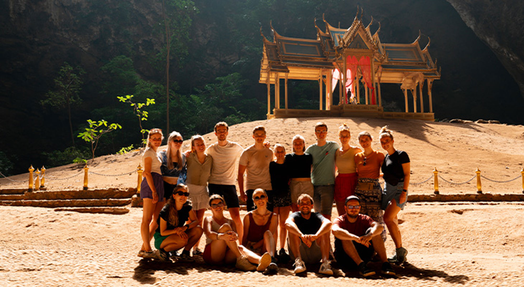 Groepsfoto van reizigers bij een Thaise tempel in een grot, met zonlicht op de ingang.