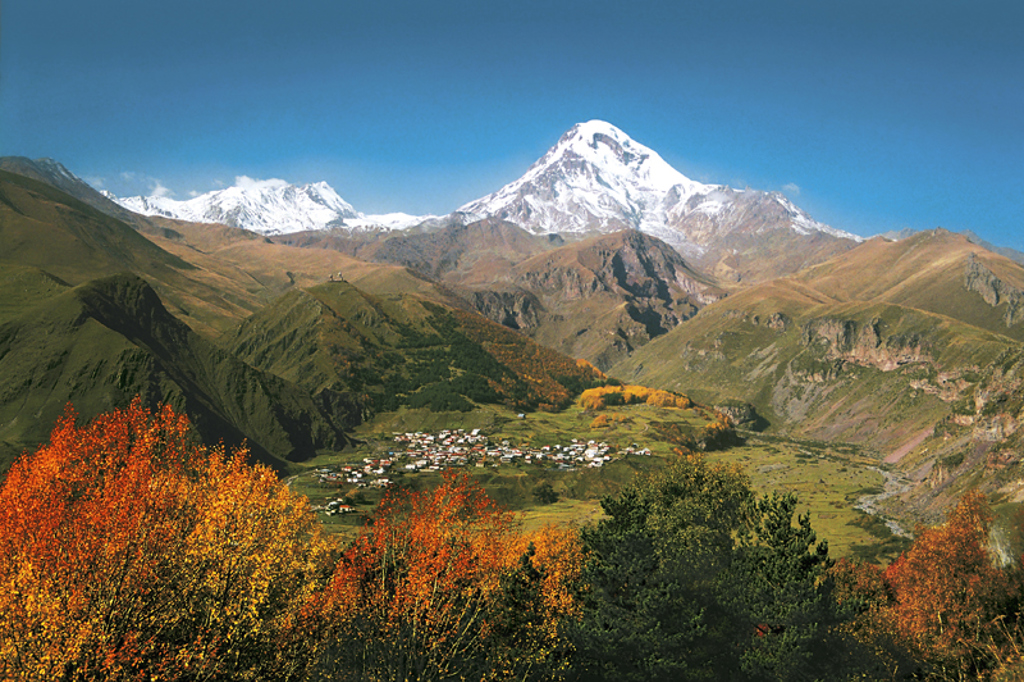 Besneeuwde bergtoppen in de Kaukasus bij Stepantsminda, met herfstkleuren en dorp in de vallei, Georgië.