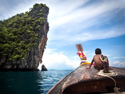 Jongen op de boeg van een longtailboot langs een hoge kalkstenen rots in de zee, Thailand.