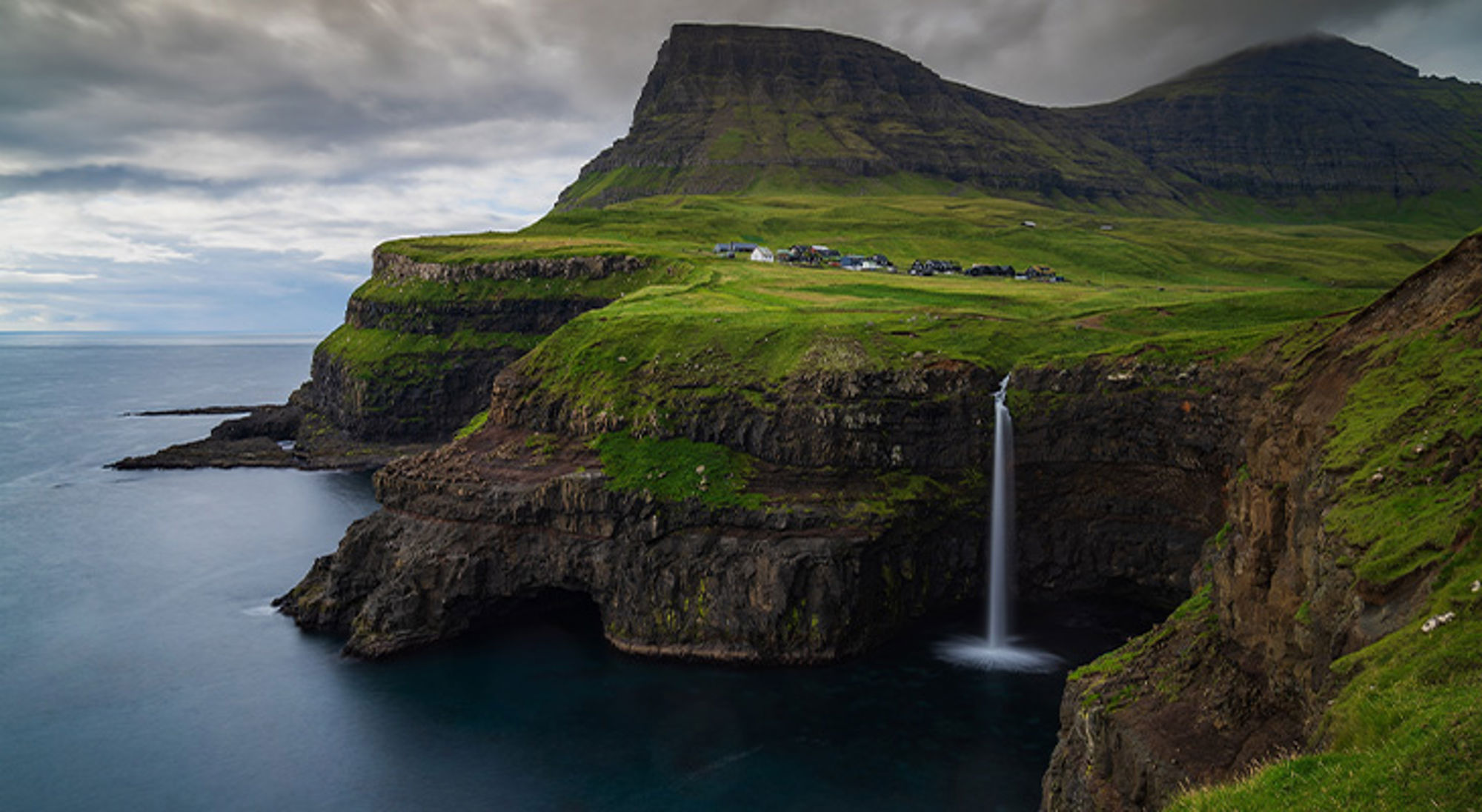 Gásadalur waterfall Groene kliffen met waterval die in zee stort, met een klein dorp op het plateau.