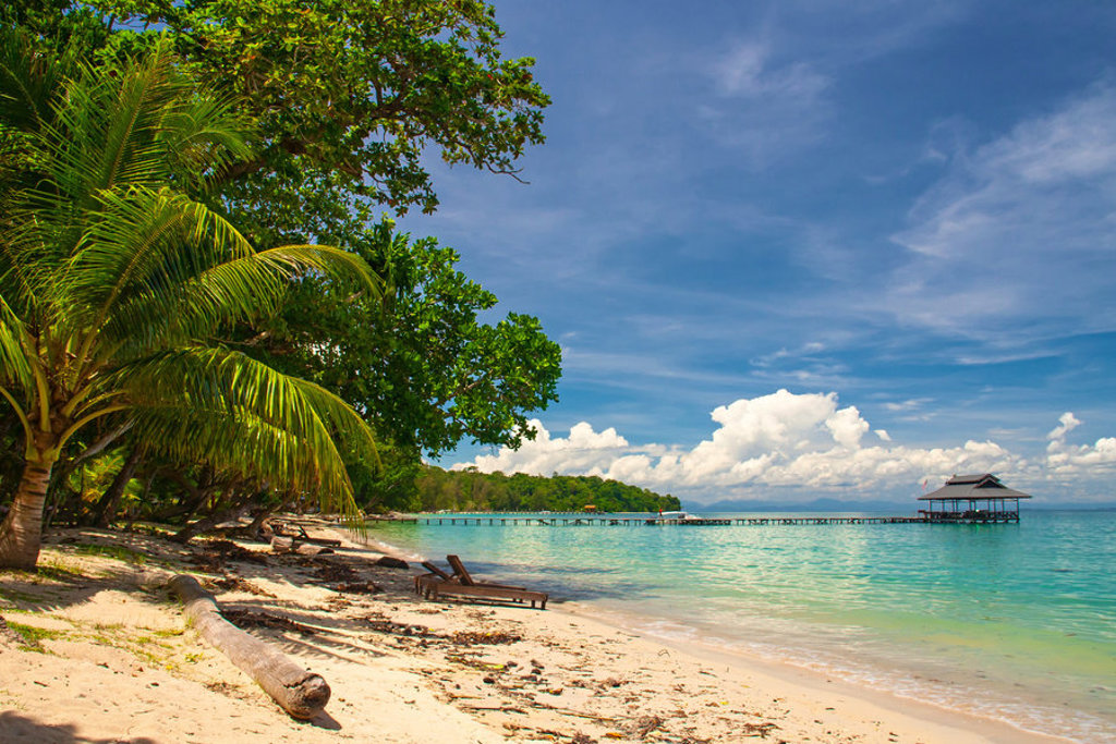 Tropisch strand in Maleisisch Borneo met palmbomen, turquoise zee en een lange pier naar een paviljoen.