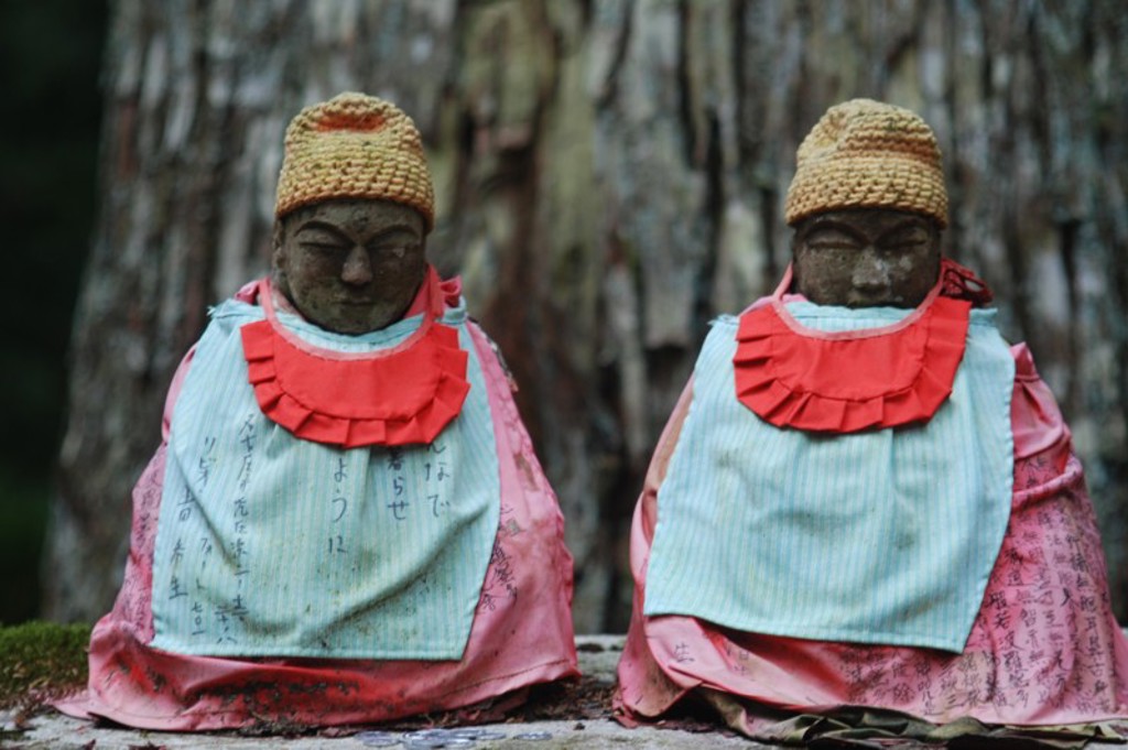 Twee Japanse Jizo-beelden met gebreide mutsen en rode slabbetjes bij een tempel tijdens een familiereis Japan.