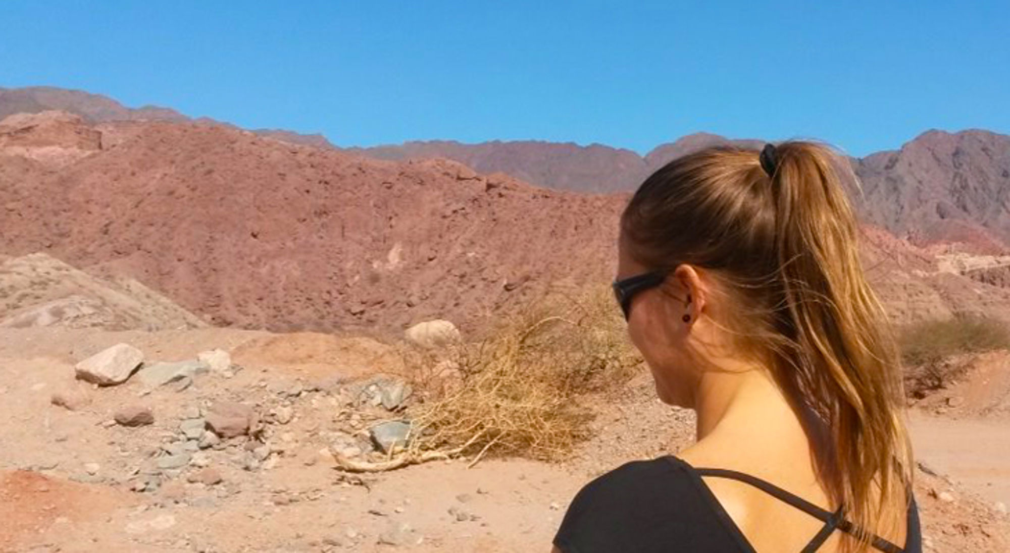 Vrouw met zonnebril kijkt uit over rotsachtige woestijnbergen onder een heldere blauwe lucht.