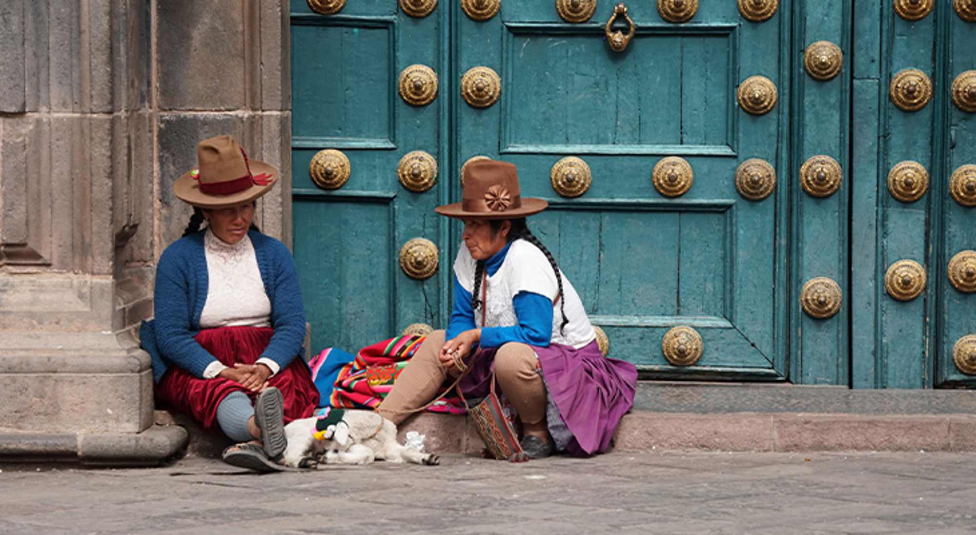 Cuzco Twee vrouwen in traditionele kleding zitten op straat voor een blauwe deur met gouden ornamenten.