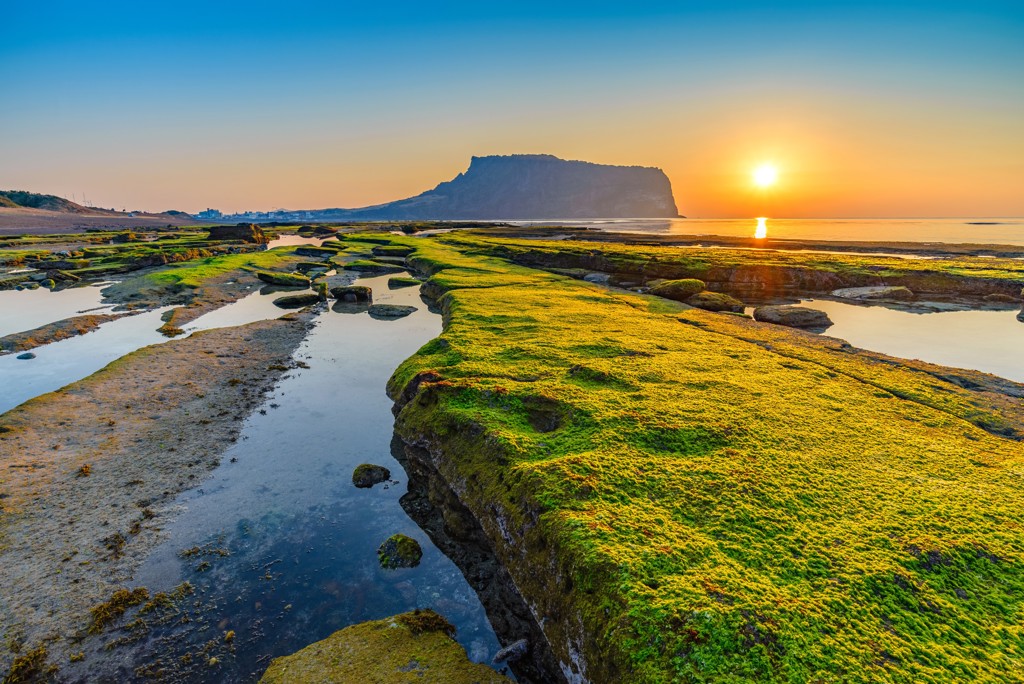 Zonsopgang bij Seongsan Ilchulbong op Jeju-eiland, met groene kustrotsen en getijdenpoelen in Zuid-Korea.