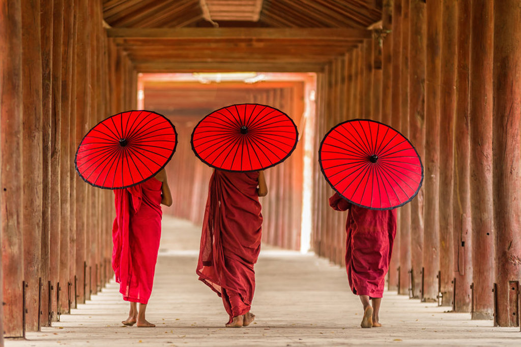 Drie boeddhistische monniken met rode parasols lopen door een houten gang in Myanmar.