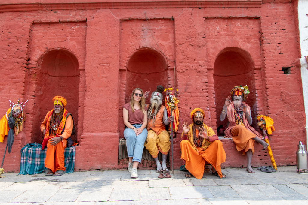 Reiziger tijdens 22-35ers reis Nepal poseert met kleurrijk geklede sadhu’s voor een rood tempelmuur.