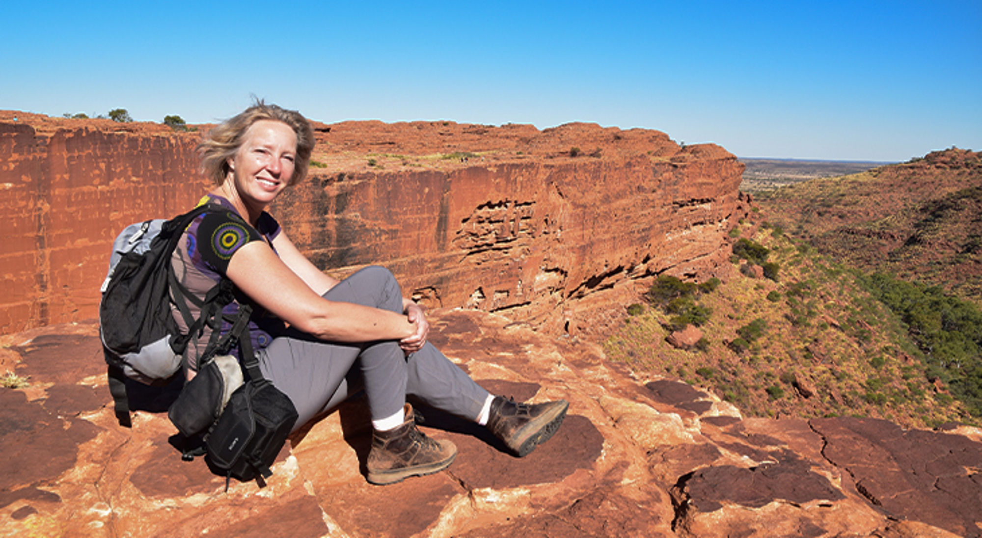 Laura Wandelaar met rugzak op rode rotsen met uitzicht op een canyon onder een blauwe lucht.