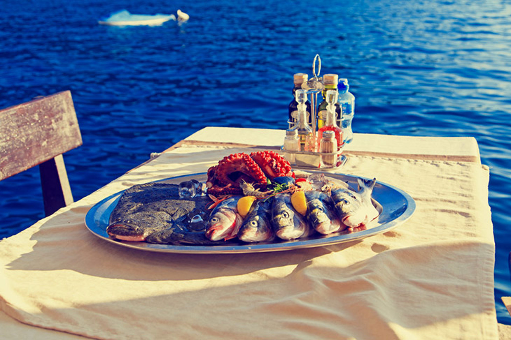 Schaal met verse vis en octopus op een tafel aan zee, met flesjes olie en kruiden op de achtergrond.