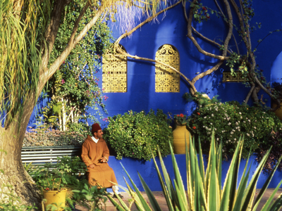 Man in traditionele kleding in de Majorelletuin in Marrakech, met felblauwe muur en gele ramen op de achtergrond.